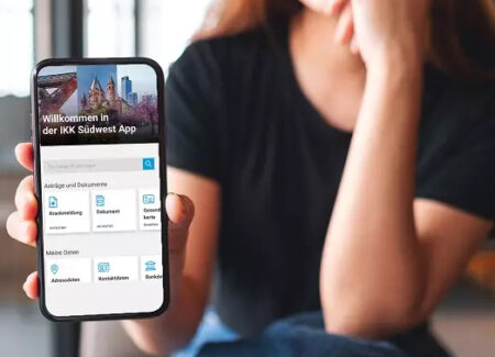 Eine Frau, die an einem Tisch sitzt, hält ein Smartphone in der Hand, auf dem der Bildschirm der "IKK Südwest App" in deutscher Sprache angezeigt wird und das verschiedene Optionen wie Dokumente, Kontaktdaten und Dienstleistungen enthält. Der Hintergrund ist sanft verschwommen.