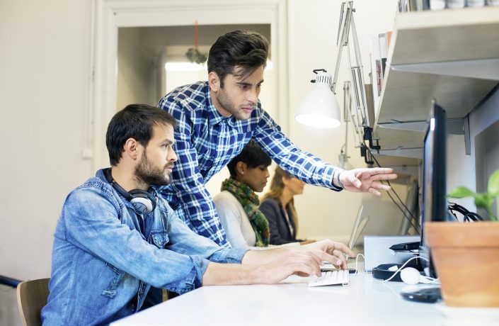 In einem Büro sitzt ein junger Mann am Schreibtisch. Ein weiterer junger Mann steht neben ihm und deutet auf den Bildschirm und erklärt ihm etwas.