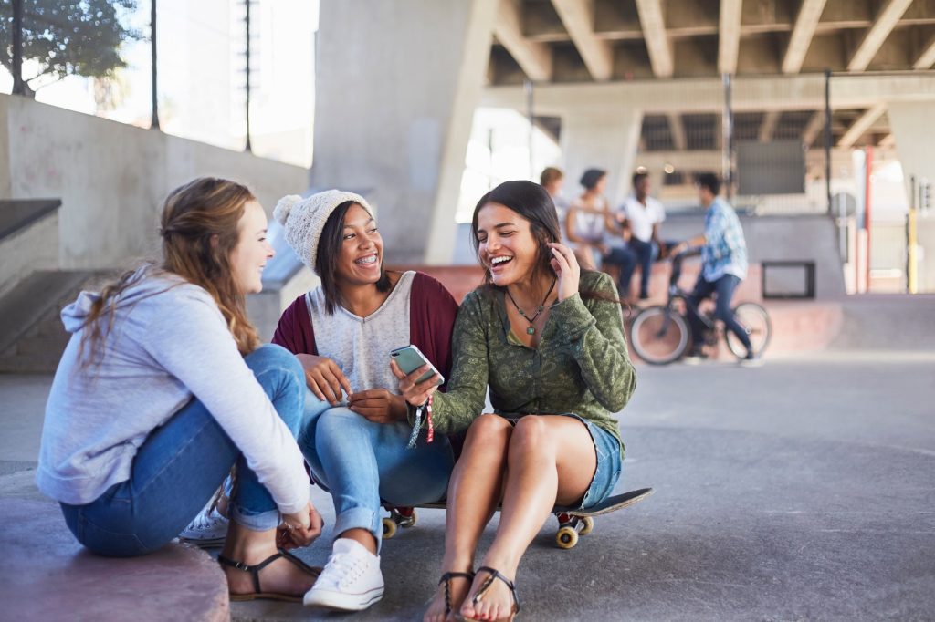 Drei Teenager sitzen gemeinsam bei einer Skateranlage und schauen gut gelaunt auf eine Skateranlage
