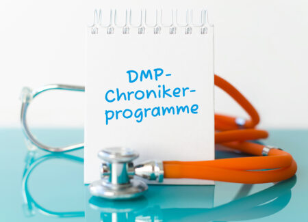 Ein Stethoskop liegt auf einer blauen Oberfläche. Daneben steht auf einem stehenden Notizzettel DMP-Chronikerprogramme.