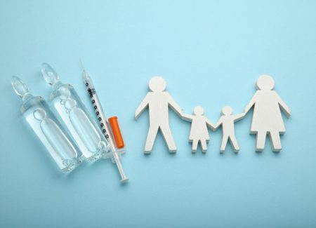Eine als Holzfigur stilisierte Familie. Daneben eine Spritze und zwei Ampullen Impfstoff.