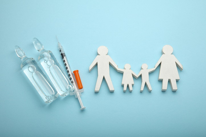 Eine als Holzfigur stilisierte Familie. Daneben eine Spritze und zwei Ampullen Impfstoff.