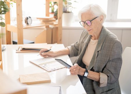 Ältere Frau sitzt am Computer und schaut auf Ihr Handy