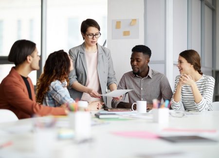 Eine Arbeitgeberin/ Professorin zeigt 4 Personen die am Tisch sitzen einen Zettel