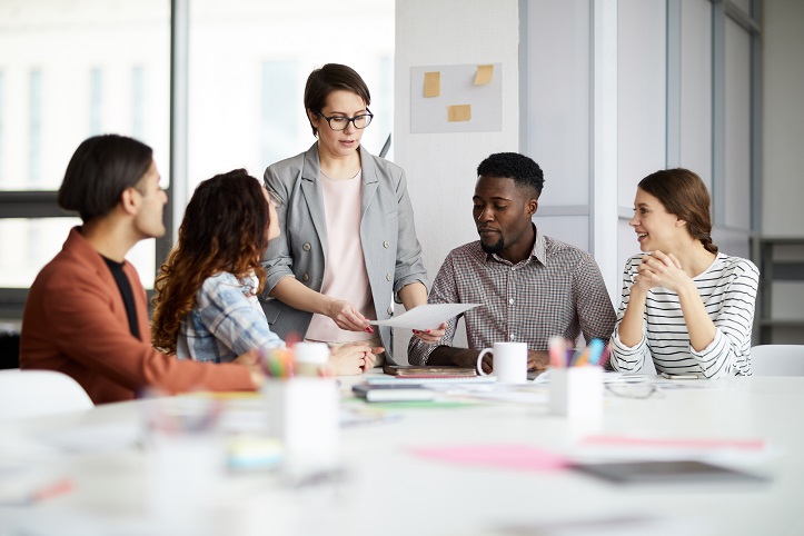 Eine Arbeitgeberin/ Professorin zeigt 4 Personen die am Tisch sitzen einen Zettel