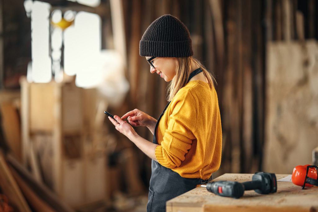 Eine junge Handwerkerin steht vor einem Tisch mit Bohrer und schaut auf ihr Smartphone