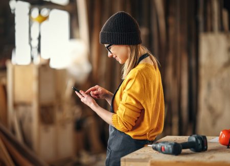 Eine junge Handwerkerin steht vor einem Tisch mit Bohrer und schaut auf ihr Smartphone