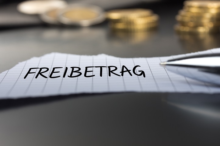 Ein Stück Zettel auf einem Schreibtisch, auf dem das Wort Freibetrag steht. Im Hintergrund sind Geldmünzen zu sehen.
