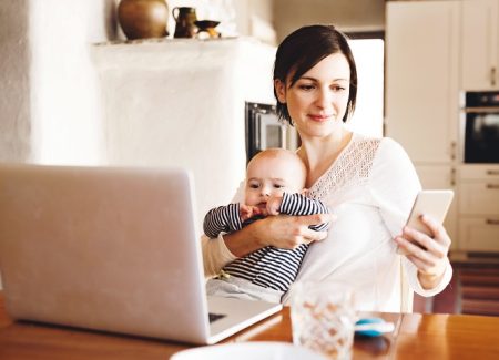 Eine junge Frau hält ein Baby im Arm, sitzt vor einem Laptop und schaut auf ein Handy