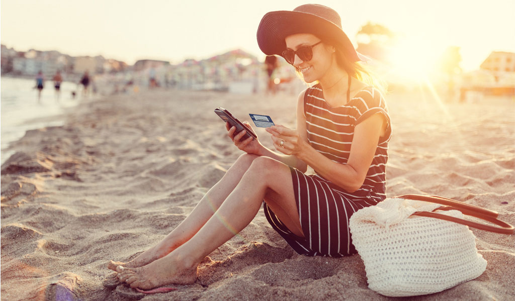 Eine Frau sitzt am Strand und schaut sowohl auf ihr Handy als auch auf die elektronische Gesundheitskarte