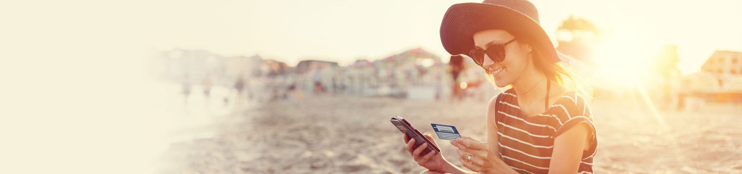 Eine Frau sitzt am Strand und schaut sowohl auf ihr Handy als auch auf die elektronische Gesundheitskarte