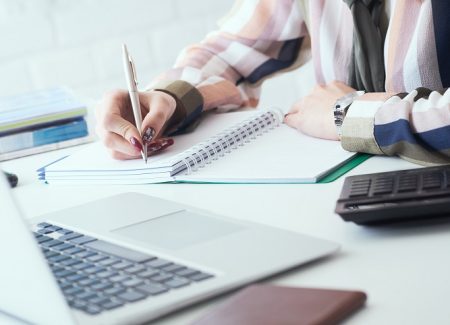 Frau sitzt vor einem Computer und schreibt auf einen Block.