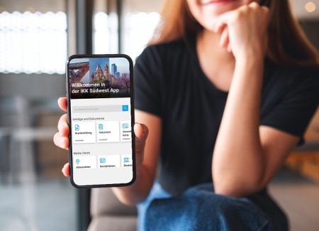 Eine junge Frau hält ein Smartphone zum Betrachter. Auf dem Display ist die Startseite der IKK Südwest-App zu sehen