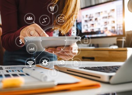 Eine Frau sitzt mit einem Tablet in der Hand vor einem Schreibtisch. Um das Tablet herum schweben Icons zum Thema Business, wie zum Beispiel Umsatz oder Warenkorb