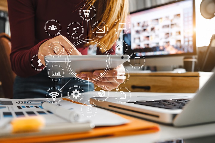 Eine Frau sitzt mit einem Tablet in der Hand vor einem Schreibtisch. Um das Tablet herum schweben Icons zum Thema Business, wie zum Beispiel Umsatz oder Warenkorb