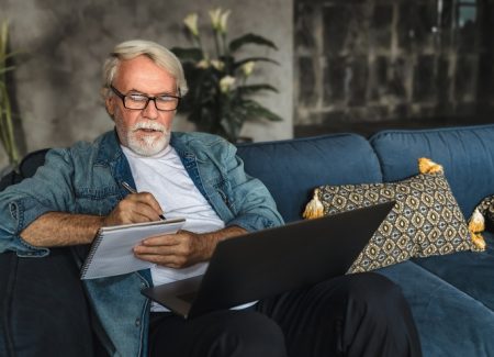 Ein Herr sitzt auf einer Couch, hat einen Notebook vor sich und macht sich Notizen in seinen Block.