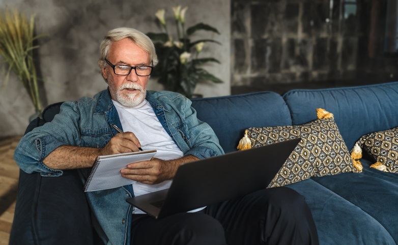 Ein Herr sitzt auf einer Couch, hat einen Notebook vor sich und macht sich Notizen in seinen Block.