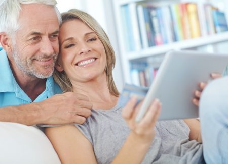 Eine Frau sitzt auf einem Sofa. Ihr Mann blickt über ihre Schulter. Gemeinsam schauen sie in ein Tablet.
