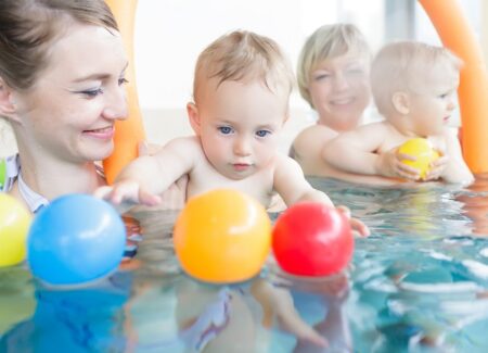Zwei Frauen in einem Schwimmbecken halten Babys, die nach bunten Bällen greifen, die auf dem Wasser schwimmen. Sowohl Babys als auch Erwachsene wirken in der hellen Schwimmhalle glücklich und beschäftigt.
