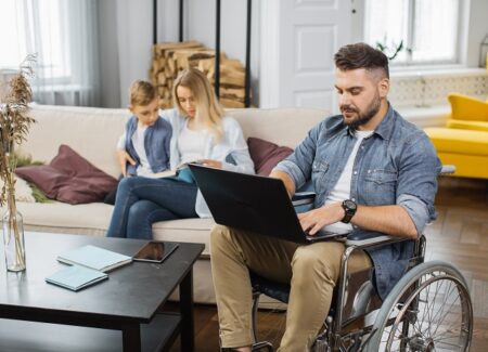 Ein Mann im Rollstuhl arbeitet am Laptop, während seine Frau und Sohn im Hintergrund auf dem Sofa sitzen