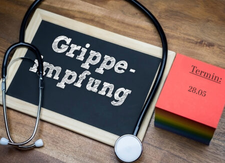Auf einer Holzfläche sind ein Stethoskop, eine Kreidetafel mit dem Wort Grippe-Impfung und ein roter Kalenderzettel mit Termin: 28.05 angeordnet.