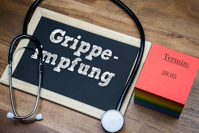 Auf einer Holzfläche sind ein Stethoskop, eine Kreidetafel mit dem Wort Grippe-Impfung und ein roter Kalenderzettel mit Termin: 28.05 angeordnet.