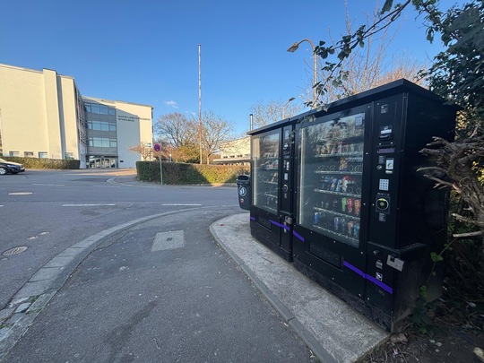 Zwei schwarze Verkaufsautomaten stehen auf einem Bürgersteig in der Nähe einer Straße, im Hintergrund ein modernes Gebäude. Der Himmel ist klar und blau, und in der Nähe gibt es einige Büsche und Bäume.