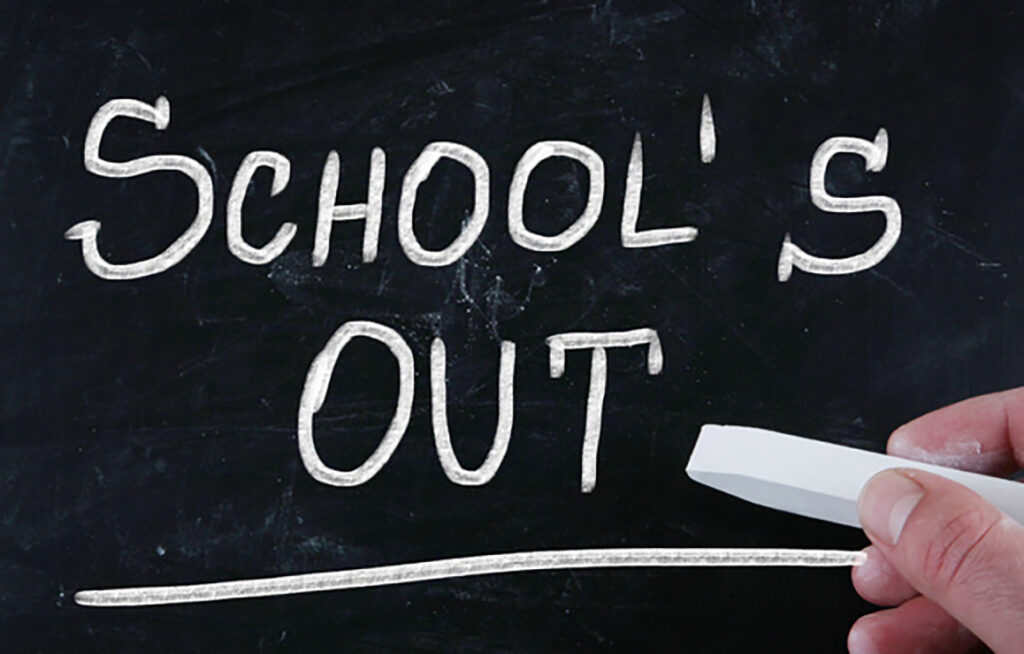 Eine Hand mit Kreide zieht einen Strich unter die in Weiß auf eine schwarze Tafel geschriebenen Worte „SCHOOLS OUT“.
