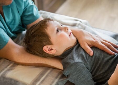 Ein kleiner Junge liegt auf dem Rücken auf einem Untersuchungstisch und blickt nach oben, während ein Erwachsener sanft eine Hand auf seine Brust und die andere Hand hinter seinen Rücken legt.