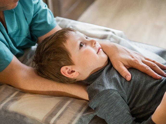 Ein kleiner Junge liegt auf dem Rücken auf einem Untersuchungstisch und blickt nach oben, während ein Erwachsener sanft eine Hand auf seine Brust und die andere Hand hinter seinen Rücken legt.
