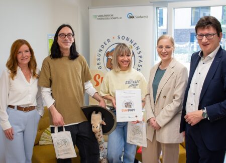 Fünf lächelnde Personen stehen in einem Büro und halten Geschenktüten und ein Zertifikat in den Händen. Hinter ihnen liegt auf einem gelben Sofa eine Plüschgiraffe. Im Hintergrund ist ein Banner mit Logos und Text zu sehen.