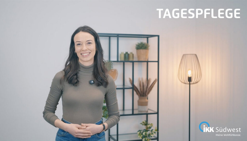 Eine Frau steht lächelnd in einem modernen Raum mit Regalen und Stehlampe. In der oberen rechten Ecke ist der Schriftzug „Tagespflege“ und in der unteren rechten Ecke das Logo der IKK Südwest zu sehen.