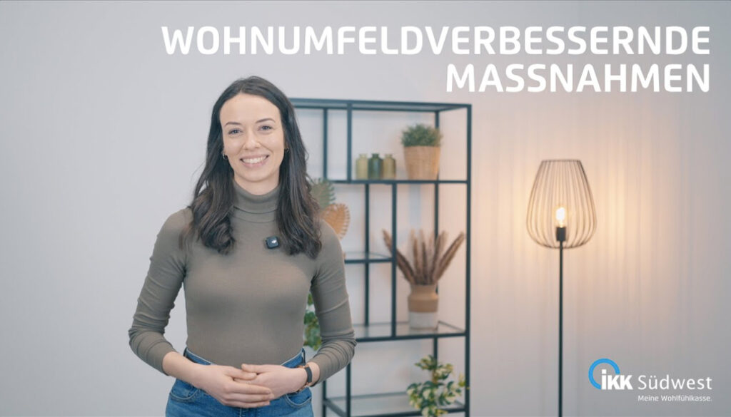 Eine Frau steht lächelnd in einem modernen Raum mit Regalen und einer Stehlampe. Über ihr steht der Text „WOHNUMFELDVERBESSERNDE MASSNAHMEN“. Unten rechts befindet sich das Logo der IKK Südwest.