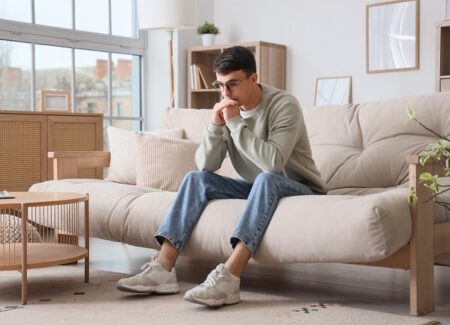 Ein junger Mann sitzt auf einem beigen Sofa in einem modernen Wohnzimmer. Er hat die Ellbogen auf den Knien und die Hände vor dem Mund und wirkt besorgt.