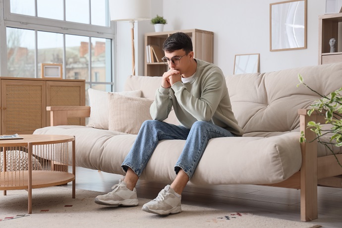 Ein junger Mann sitzt auf einem beigen Sofa in einem modernen Wohnzimmer. Er hat die Ellbogen auf den Knien und die Hände vor dem Mund und wirkt besorgt.