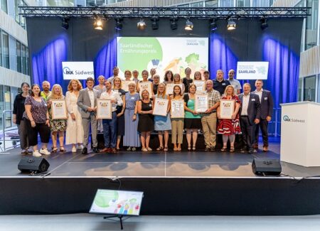 Eine Gruppe von Personen steht auf einer Bühne und posiert für ein Foto bei einer Preisverleihung. Mehrere Personen halten gerahmte Urkunden in der Hand. Hinter ihnen sind Veranstaltungsbanner, helle Bühnenbeleuchtung und ein Podium mit der Aufschrift IKK Südwest zu sehen.