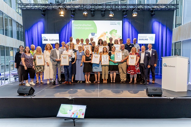 Eine Gruppe von Personen steht auf einer Bühne und posiert für ein Foto bei einer Preisverleihung. Mehrere Personen halten gerahmte Urkunden in der Hand. Hinter ihnen sind Veranstaltungsbanner, helle Bühnenbeleuchtung und ein Podium mit der Aufschrift IKK Südwest zu sehen.
