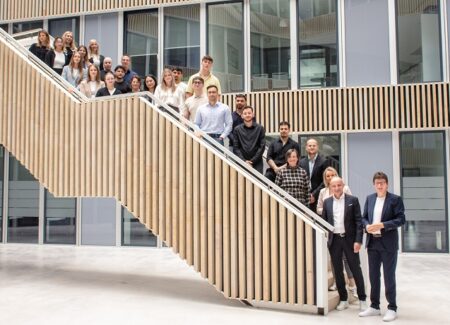 Eine Gruppe von fünfzehn Personen, gekleidet in Geschäfts- und Freizeitkleidung, steht in einem modernen, hellen Atrium mit Glaswänden und einer breiten Treppe im Hintergrund.