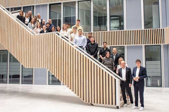Eine Gruppe von fünfzehn Personen, gekleidet in Geschäfts- und Freizeitkleidung, steht in einem modernen, hellen Atrium mit Glaswänden und einer breiten Treppe im Hintergrund.