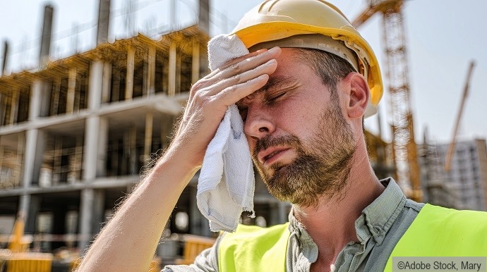 Ein Bauarbeiter, der einen gelben Schutzhelm und eine Sicherheitsweste trägt, wischt sich mit einem Tuch den Schweiß von der Stirn und sieht müde aus, während im Hintergrund ein unfertiges Gebäude zu sehen ist.