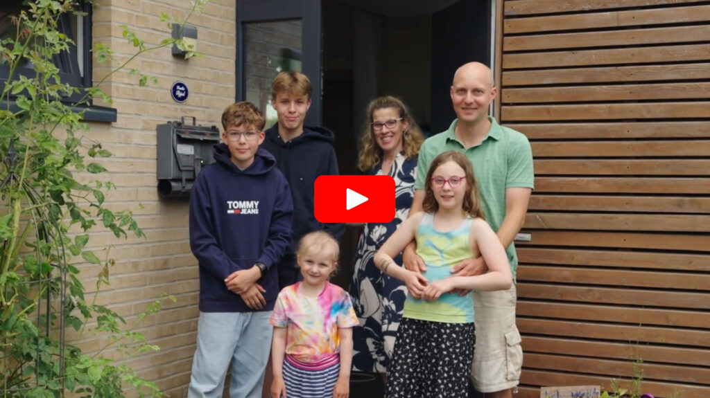 Eine sechsköpfige Familie, darunter zwei Erwachsene und vier Kinder, steht lächelnd vor dem Eingang eines modernen Hauses mit Holzfassade und Pflanzen in der Nähe.