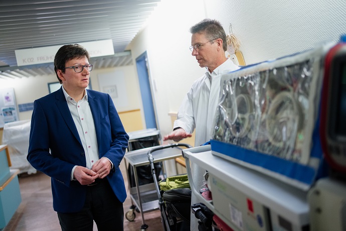 Zwei Männer stehen in einem Krankenhauskorridor. Der Vorstand der IKK Südwest trägt einen blauen Anzug und hört zu, während der Arzt in einem weißen Laborkittel, in Richtung medizinischer Geräte gestikuliert. Im Vordergrund sind ein medizinischer Wagen und ein Inkubator zu sehen.