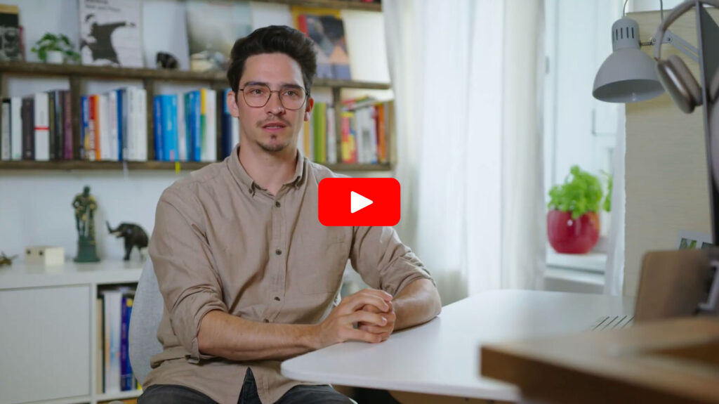 Ein Mann mit Brille und beigem Hemd sitzt an einem weißen Schreibtisch in einem hellen Raum mit Bücherregalen, einer Pflanze und einem Fenster im Hintergrund. Er blickt nach vorne und stützt seine Hände auf den Schreibtisch.