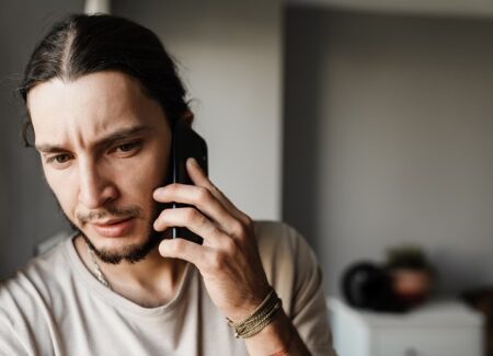 Ein junger Mann mit langen Haaren und Bart schaut besorgt, während er in einem Haus mit einem Smartphone spricht. Er trägt ein helles Hemd und Armbänder am Handgelenk.