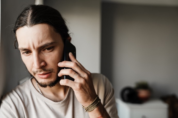 Ein junger Mann mit langen Haaren und Bart schaut besorgt, während er in einem Haus mit einem Smartphone spricht. Er trägt ein helles Hemd und Armbänder am Handgelenk.