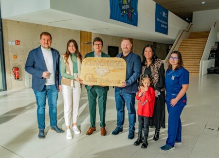 Sieben Personen stehen in einem Innenraum, lächeln und halten ein Holzschild mit der Aufschrift "Kinderlachen Stiftung" und "Saar Südwest" mit einer Handschlaggrafik. Ein Kind ist in der Gruppe und im Hintergrund ist eine große Treppe zu sehen.