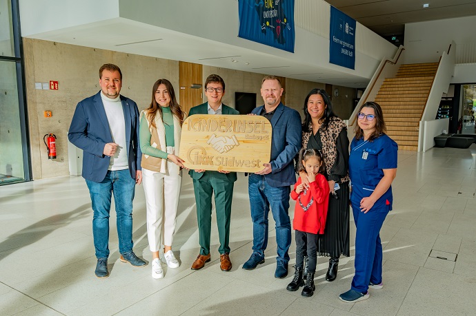 Sieben Personen stehen in einem Innenraum, lächeln und halten ein Holzschild mit der Aufschrift "Kinderlachen Stiftung" und "Saar Südwest" mit einer Handschlaggrafik. Ein Kind ist in der Gruppe und im Hintergrund ist eine große Treppe zu sehen.