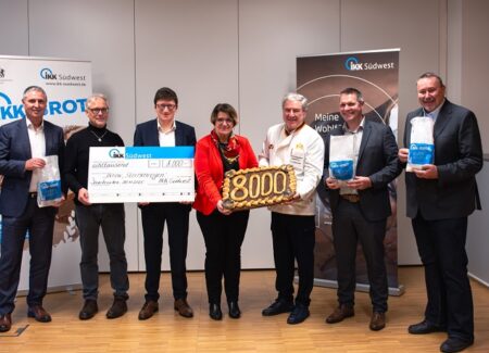 Sieben Personen stehen in einem Raum und halten einen großen Scheck, einen 8000er Kuchen und Informationsbroschüren in der Hand. Im Hintergrund sind zwei Banner mit den Logos der IKK Südwest zu sehen.
