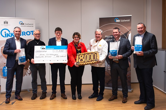 Sieben Personen stehen in einem Raum und halten einen großen Scheck, einen 8000er Kuchen und Informationsbroschüren in der Hand. Im Hintergrund sind zwei Banner mit den Logos der IKK Südwest zu sehen.