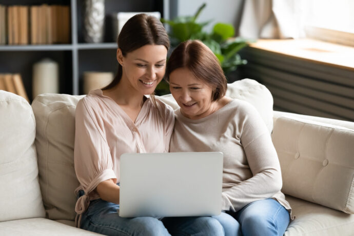 Zwei Frauen sitzen eng beieinander auf einer Couch und schauen lächelnd auf einen Laptop-Bildschirm.